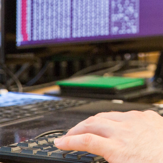 hand typing on a keyboard with a computer screen displaying code in the background
