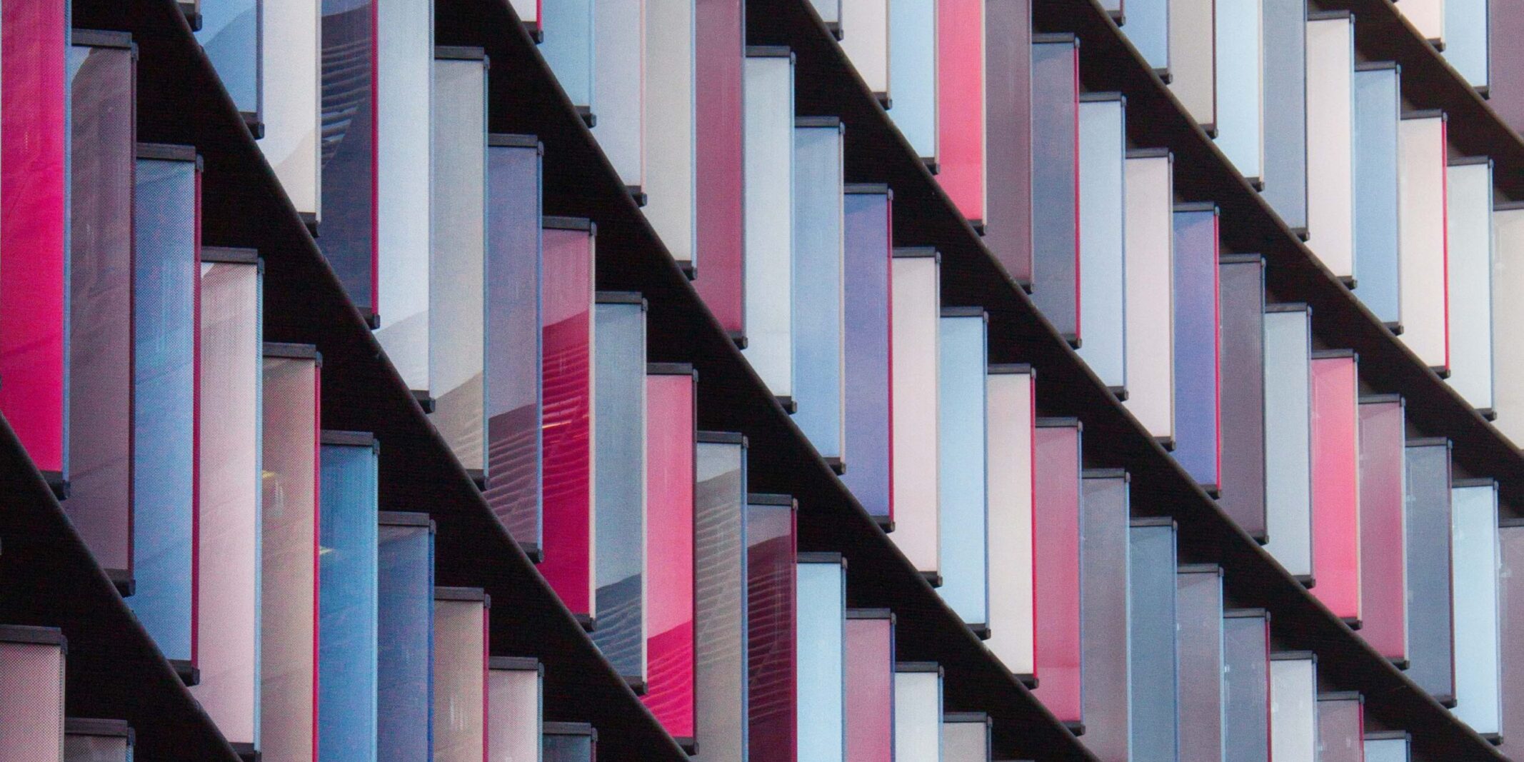 Modern building facade with repeating rows of glass panels in shades of blue, white, and pink arranged in a diagonal pattern
