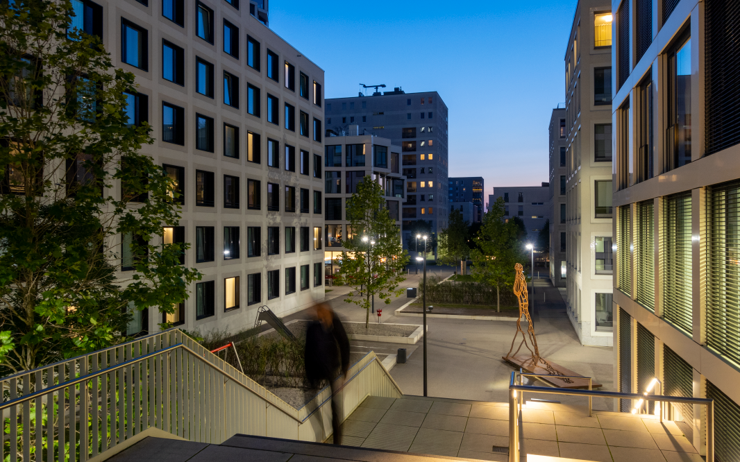 Modern apartment buildings surrounding a courtyard at dusk, with lit windows, trees, a staircase in the foreground, and a metal sculpture in the open space