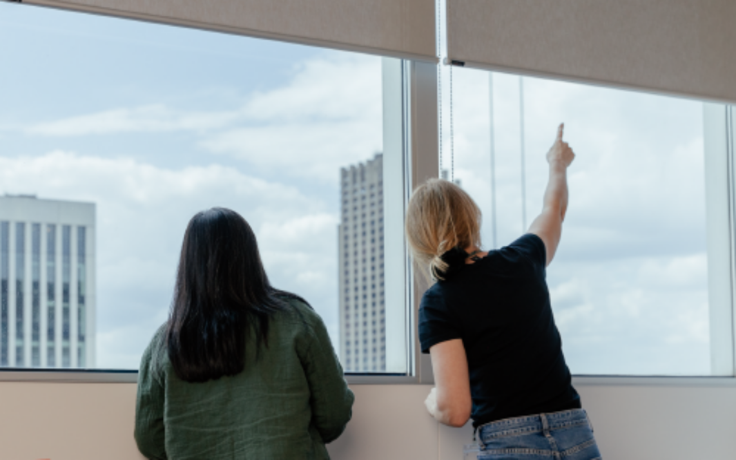 Two people standing by a window looking outside at city buildings, with one person pointing upward toward the sky