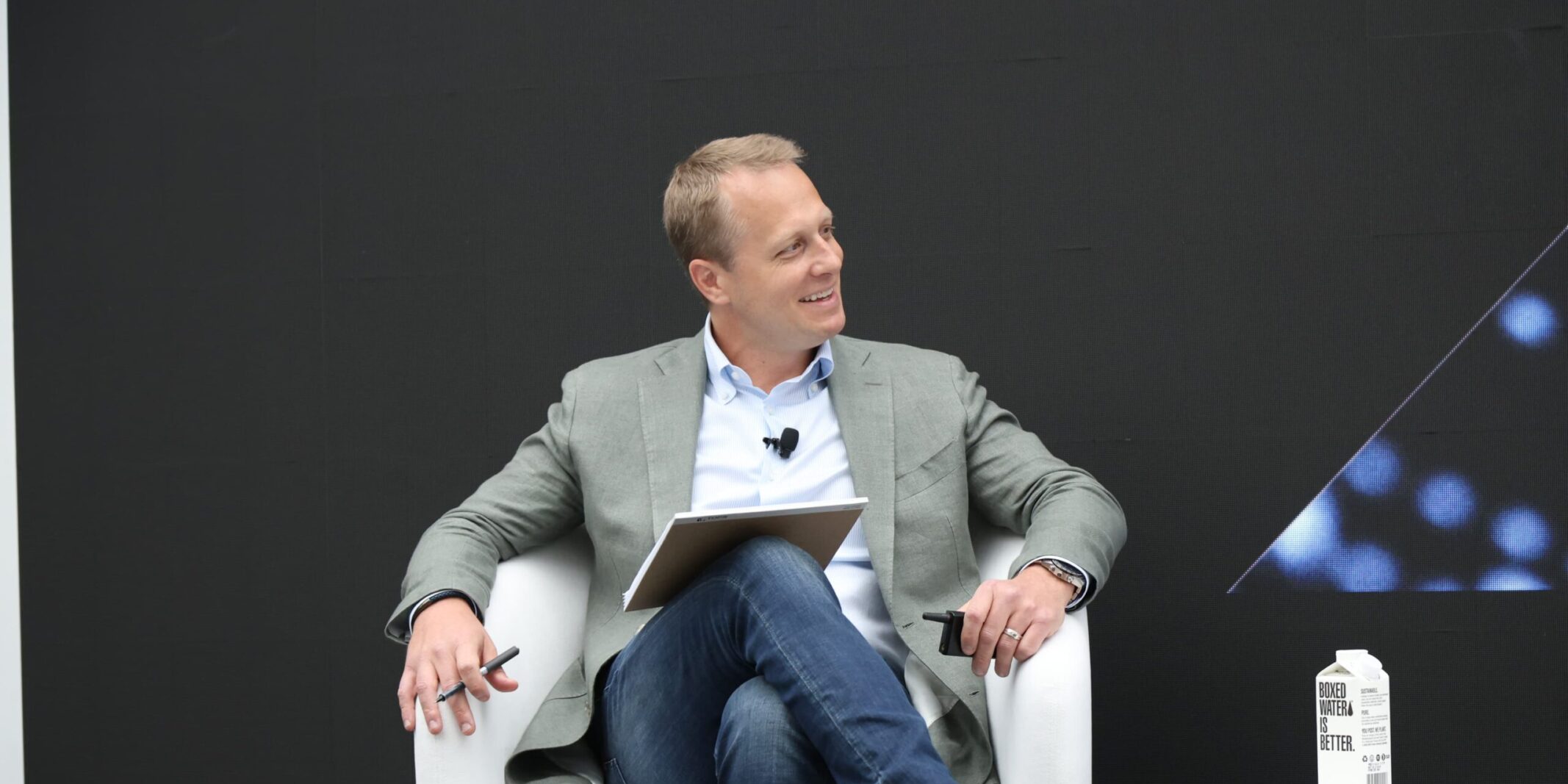 A man in a light green blazer and jeans sits on a white chair on stage, holding a pen and tablet while smiling and looking to the side, with a small table and a boxed water container beside him against a dark background